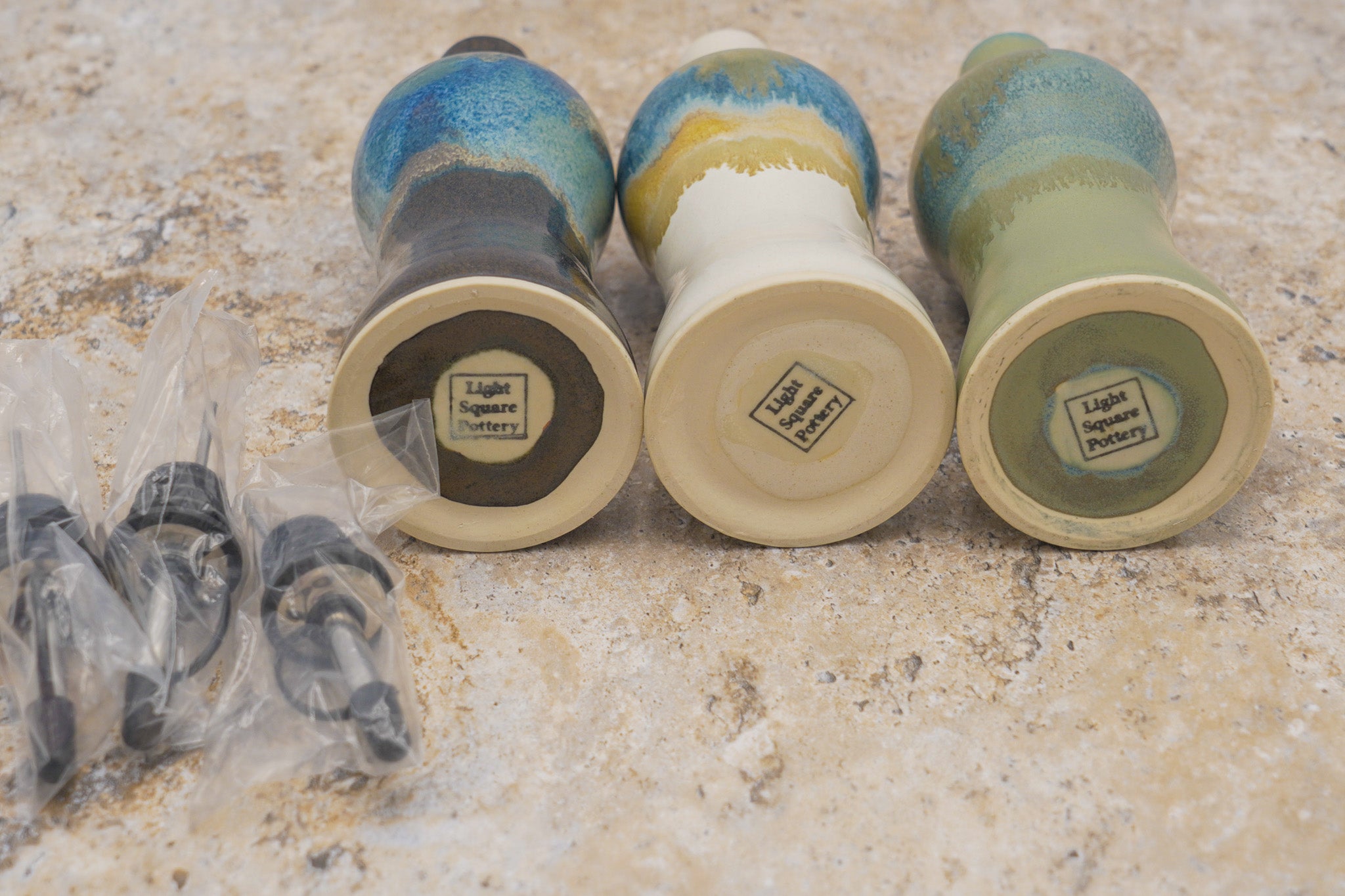 Bottom view of 3 ceramic pourer bottles with different color glazes on a marble surface, wrapped new pour tops sitting next to them, glazed over maker's mark stamp saying Light Square Pottery on bottoms